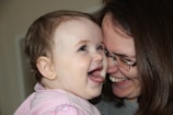 A baby and an adult sharing a joyful moment, both smiling and expressing happiness. The baby is wearing a pink outfit and seems to be looking towards the adult, possibly their mother, who is wearing glasses.