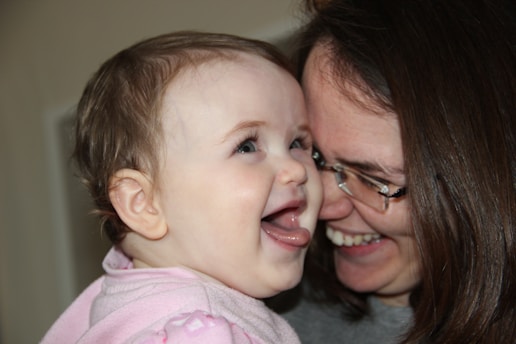 A baby and an adult sharing a joyful moment, both smiling and expressing happiness. The baby is wearing a pink outfit and seems to be looking towards the adult, possibly their mother, who is wearing glasses.