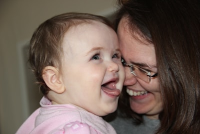 A baby and an adult sharing a joyful moment, both smiling and expressing happiness. The baby is wearing a pink outfit and seems to be looking towards the adult, possibly their mother, who is wearing glasses.