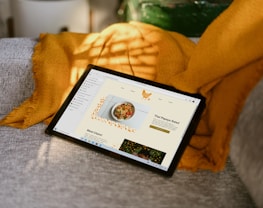 A tablet placed on a gray couch, partially covered by a mustard yellow blanket. The tablet screen displays a webpage featuring a recipe for Thai papaya salad, with a vibrant image of the dish and decorative elements. Sunlight casts a pattern of shadows across the scene, adding a warm and cozy ambiance.