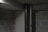 A concrete and cinder block basement room with two vertical pipes, one metallic and the other dark in color. The area is dimly lit, casting shadows on the walls, and there's a red box affixed to the wall.