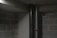 A concrete and cinder block basement room with two vertical pipes, one metallic and the other dark in color. The area is dimly lit, casting shadows on the walls, and there's a red box affixed to the wall.