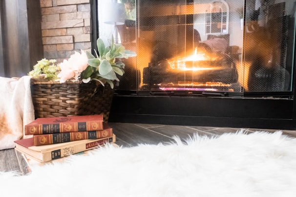 A cozy living space with a lit fireplace that casts a warm glow. A basket with flowers and green foliage sits beside a stack of vintage books. The room features a fluffy white rug and a stone accent wall.