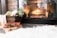 A cozy living space with a lit fireplace that casts a warm glow. A basket with flowers and green foliage sits beside a stack of vintage books. The room features a fluffy white rug and a stone accent wall.