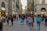 A bustling pedestrian street is lined with shops and historic buildings. Groups of people walk in various directions, carrying bags and wearing casual attire. The architecture features arched windows and ornate details typical of European city centers.