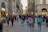 A bustling pedestrian street is lined with shops and historic buildings. Groups of people walk in various directions, carrying bags and wearing casual attire. The architecture features arched windows and ornate details typical of European city centers.