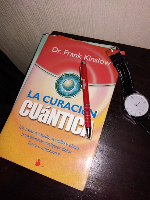 A book titled 'La Curación Cuántica' by Dr. Frank Kinslow is lying on a dark wooden surface. On top of the book is a red pen and next to it is a wristwatch with a black leather strap.