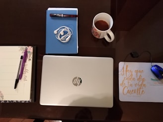 A neatly arranged desk setup includes a closed silver laptop, a notebook with a pen, a pair of tangled earphones, a mug with a drink, a planner with a pen, and a mouse on a pad. The mouse pad has motivational text written on it.
