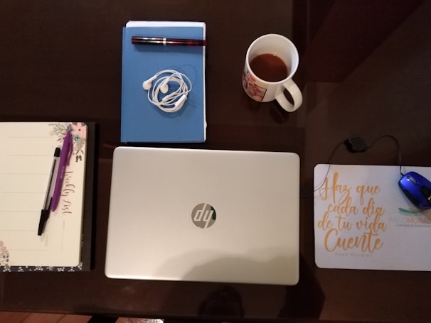 A neatly arranged desk setup includes a closed silver laptop, a notebook with a pen, a pair of tangled earphones, a mug with a drink, a planner with a pen, and a mouse on a pad. The mouse pad has motivational text written on it.