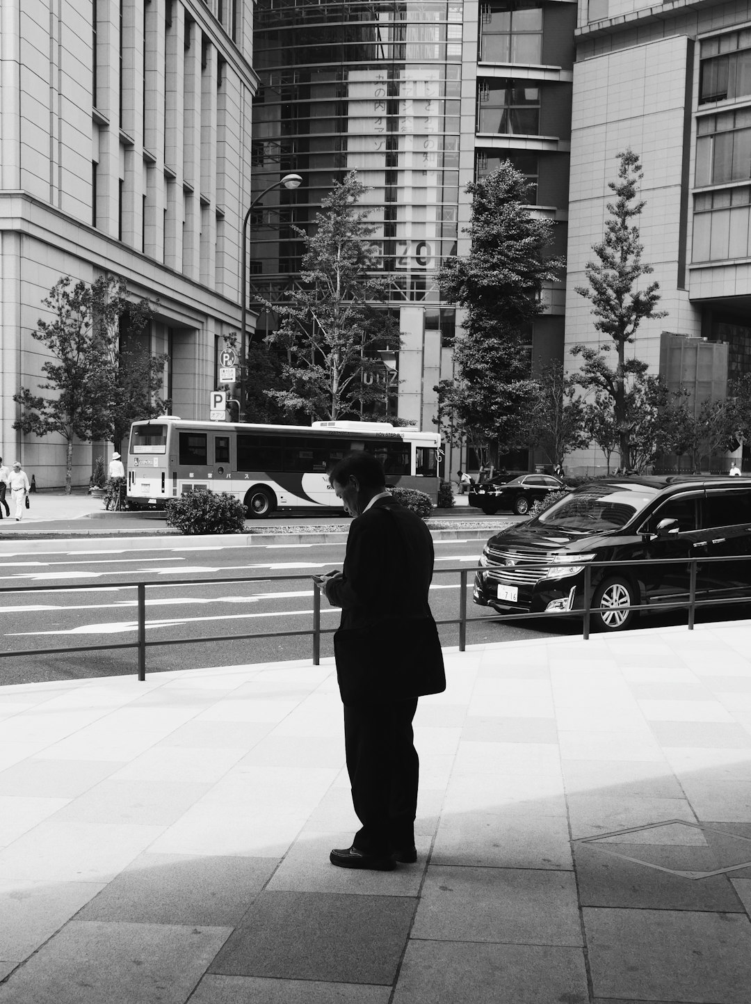 grayscale photo of man in black suit standing on sidewalk near cars and buildings