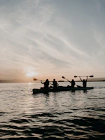 A group of friends paddling kayaks on calm blue waters at sunset.