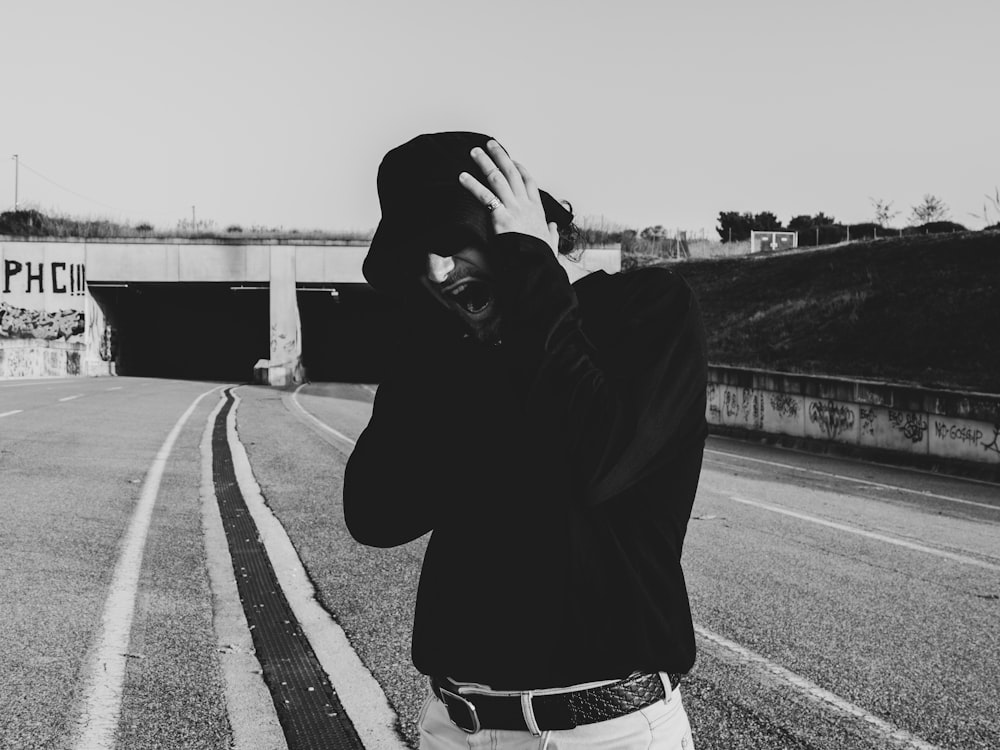 grayscale photo of man in black hoodie and white pants standing on road