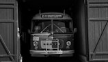 A vintage Volvo bus is parked in a dimly lit garage with large wooden doors partially open on both sides. The bus is well-preserved, showcasing a classic design with rounded edges, large headlights, and a front-mounted grille. The signage above the windshield displays route details.
