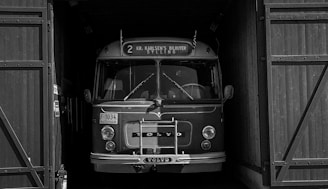A vintage Volvo bus is parked in a dimly lit garage with large wooden doors partially open on both sides. The bus is well-preserved, showcasing a classic design with rounded edges, large headlights, and a front-mounted grille. The signage above the windshield displays route details.