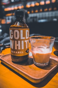 A bottle labeled 'Cold White Caramel' and a glass containing a caramel-colored beverage are placed on a wooden tray. The scene appears to be in a cozy, warmly lit setting with blurred background elements suggesting a café or bar environment.