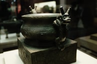 An intricately designed ancient bronze vessel with detailed engravings and a sculpted animal figure as part of the handle. The vessel rests on a pedestal with similar designs in a museum setting.