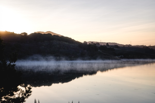 A serene lake with a gentle mist hovering over its surface during sunrise. The landscape features a calm body of water reflecting the silhouette of surrounding hills, while the sky in the background displays soft pastel hues. Trees and bushes frame the foreground, creating a natural and tranquil scene.