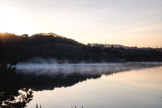 A serene lake with a gentle mist hovering over its surface during sunrise. The landscape features a calm body of water reflecting the silhouette of surrounding hills, while the sky in the background displays soft pastel hues. Trees and bushes frame the foreground, creating a natural and tranquil scene.