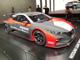 A sleek, sporty Nissan car featuring a dynamic design is displayed in a well-lit showroom. The vehicle has prominent branding with 'Nissan Intelligent Mobility' and 'Nismo' logos. It showcases a blend of red, white, and gray colors with aerodynamic features such as a spoiler. Several people are looking at the car, indicating a possible event or car show.
