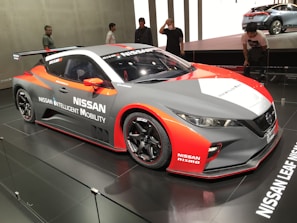 A sleek, sporty Nissan car featuring a dynamic design is displayed in a well-lit showroom. The vehicle has prominent branding with 'Nissan Intelligent Mobility' and 'Nismo' logos. It showcases a blend of red, white, and gray colors with aerodynamic features such as a spoiler. Several people are looking at the car, indicating a possible event or car show.
