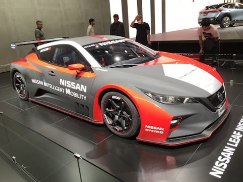 A sleek, sporty Nissan car featuring a dynamic design is displayed in a well-lit showroom. The vehicle has prominent branding with 'Nissan Intelligent Mobility' and 'Nismo' logos. It showcases a blend of red, white, and gray colors with aerodynamic features such as a spoiler. Several people are looking at the car, indicating a possible event or car show.