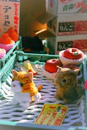 A market stall with plush toys, including a small cat and an owl, in front of a display containing packaged apples and oranges. Bright lighting illuminates the scene, creating a warm and inviting atmosphere. Japanese text appears on boxes and a yellow price tag, adding a cultural context to the setting.