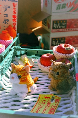 A market stall with plush toys, including a small cat and an owl, in front of a display containing packaged apples and oranges. Bright lighting illuminates the scene, creating a warm and inviting atmosphere. Japanese text appears on boxes and a yellow price tag, adding a cultural context to the setting.