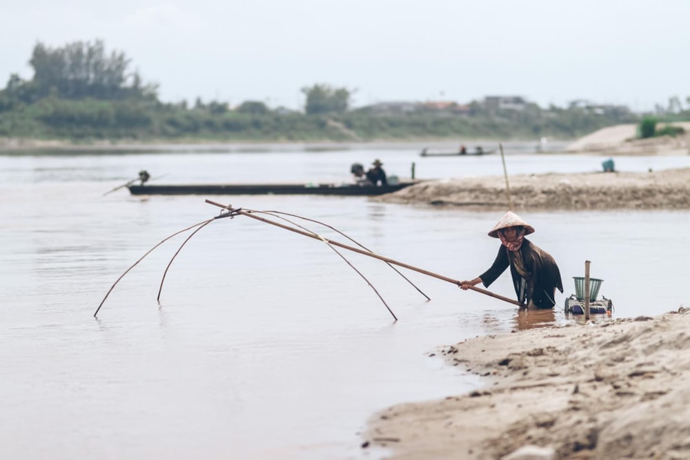 Femme en chemise bleue et chapeau marron tenant un bâton brun près d’un plan d’eau pendant la journée