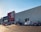 A large commercial retail store with a prominent red and white logo on the facade. Several cars are parked in the lot in front of the building, and a person is walking towards the entrance under a clear blue sky.