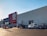A large commercial retail store with a prominent red and white logo on the facade. Several cars are parked in the lot in front of the building, and a person is walking towards the entrance under a clear blue sky.