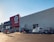 A large commercial retail store with a prominent red and white logo on the facade. Several cars are parked in the lot in front of the building, and a person is walking towards the entrance under a clear blue sky.