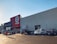 A large commercial retail store with a prominent red and white logo on the facade. Several cars are parked in the lot in front of the building, and a person is walking towards the entrance under a clear blue sky.