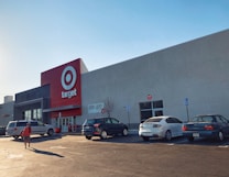 A large commercial retail store with a prominent red and white logo on the facade. Several cars are parked in the lot in front of the building, and a person is walking towards the entrance under a clear blue sky.