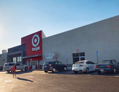 A large commercial retail store with a prominent red and white logo on the facade. Several cars are parked in the lot in front of the building, and a person is walking towards the entrance under a clear blue sky.