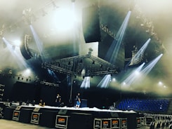 A large stage setup in an indoor arena with several people working on it. The stage is equipped with multiple lighting fixtures and large speaker systems. The ceiling features complex rigging and lighting structures. Blue seats are visible in the background, suggesting the venue is ready for a performance.