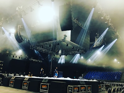 A large stage setup in an indoor arena with several people working on it. The stage is equipped with multiple lighting fixtures and large speaker systems. The ceiling features complex rigging and lighting structures. Blue seats are visible in the background, suggesting the venue is ready for a performance.