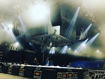 A large stage setup in an indoor arena with several people working on it. The stage is equipped with multiple lighting fixtures and large speaker systems. The ceiling features complex rigging and lighting structures. Blue seats are visible in the background, suggesting the venue is ready for a performance.