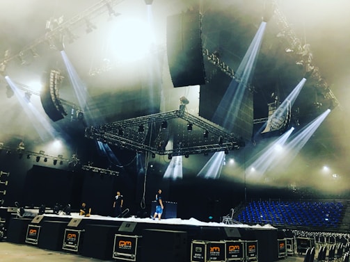 A large stage setup in an indoor arena with several people working on it. The stage is equipped with multiple lighting fixtures and large speaker systems. The ceiling features complex rigging and lighting structures. Blue seats are visible in the background, suggesting the venue is ready for a performance.
