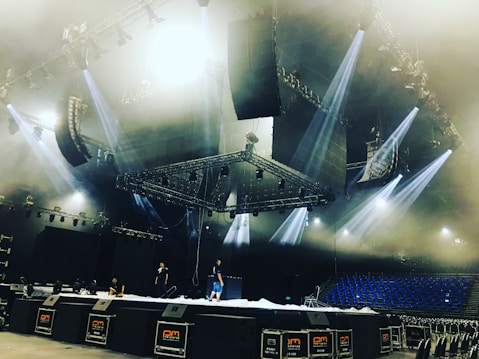 A large stage setup in an indoor arena with several people working on it. The stage is equipped with multiple lighting fixtures and large speaker systems. The ceiling features complex rigging and lighting structures. Blue seats are visible in the background, suggesting the venue is ready for a performance.