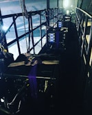 A narrow, industrial-looking platform with a series of large, black equipment cases secured with straps. Several control panels with glowing blue lights are visible on the front of each case. The platform is enclosed with metal railings, and various cables and chains hang along the sides, adding to the technical and utilitarian atmosphere. Bright lighting from behind casts a dramatic contrast against the darker sections of the platform.