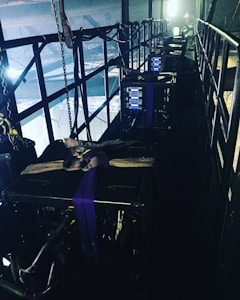 A narrow, industrial-looking platform with a series of large, black equipment cases secured with straps. Several control panels with glowing blue lights are visible on the front of each case. The platform is enclosed with metal railings, and various cables and chains hang along the sides, adding to the technical and utilitarian atmosphere. Bright lighting from behind casts a dramatic contrast against the darker sections of the platform.