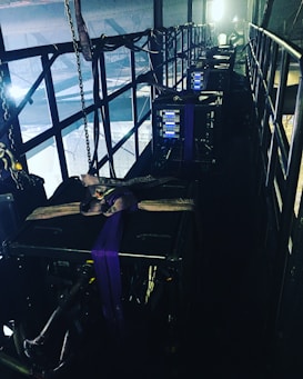 A narrow, industrial-looking platform with a series of large, black equipment cases secured with straps. Several control panels with glowing blue lights are visible on the front of each case. The platform is enclosed with metal railings, and various cables and chains hang along the sides, adding to the technical and utilitarian atmosphere. Bright lighting from behind casts a dramatic contrast against the darker sections of the platform.