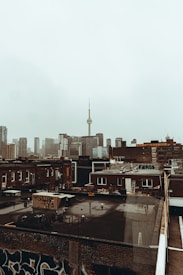 Urban cityscape featuring a cluster of high-rise buildings under a gray, overcast sky. The foreground shows older buildings with graffiti on the walls and rooftops, contrasting with the modern skyscrapers in the background. The prominent CN Tower is visible, extending into the sky, serving as a focal point.