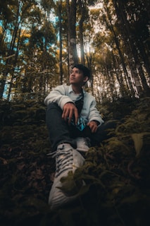 A young person sits in a forest, surrounded by tall trees and lush greenery. They wear a casual outfit, including a white jacket and sneakers, and look contemplatively into the distance. Sunlight filters through the dense canopy above, creating a serene and natural atmosphere.