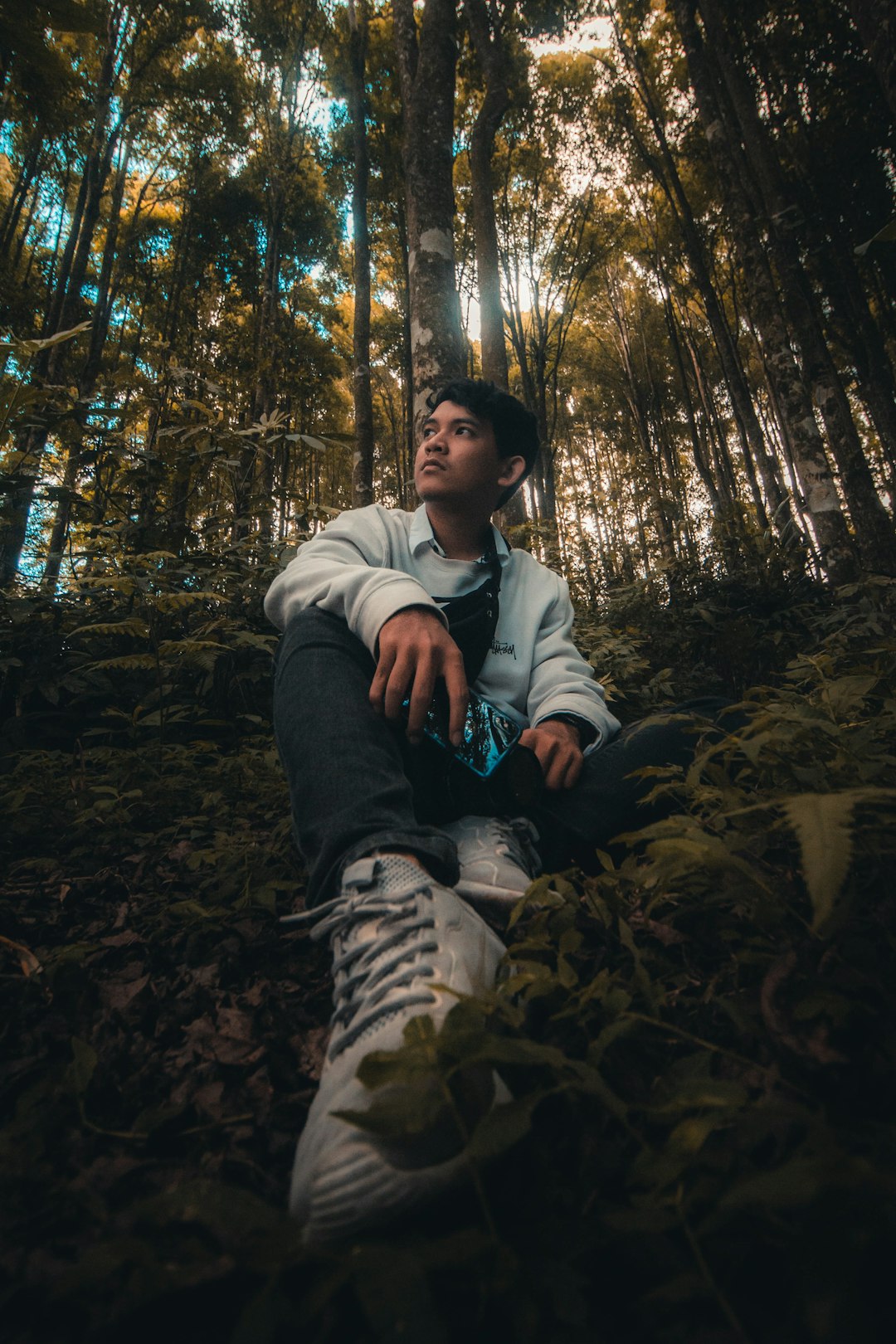 man in white hoodie sitting on ground surrounded by trees during daytime