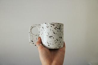 A hand is holding a white ceramic mug with black speckles. The mug has a modern, chunky handle design and is set against a plain, neutral background.