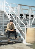 A person is sitting on an outdoor metal staircase, holding a remote control while observing a hovering drone nearby. The surroundings appear industrial with metal structures and railings under a clear blue sky.