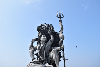 A large statue of a deity with intricate details is situated outdoors. The figure holds a trident and is adorned with various ornaments and garlands. The sky is clear and blue, with a lone bird flying nearby.