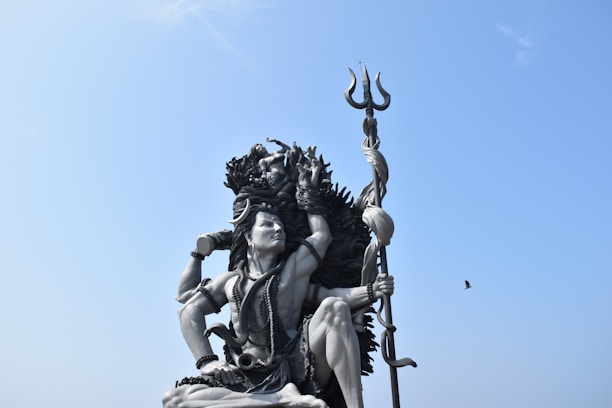 A large statue of a deity with intricate details is situated outdoors. The figure holds a trident and is adorned with various ornaments and garlands. The sky is clear and blue, with a lone bird flying nearby.