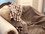 A plush, ornately carved sofa is draped with a patterned, brown and white checked blanket. A soft, floral-patterned pillow in gold hues complements the luxury of the setting.